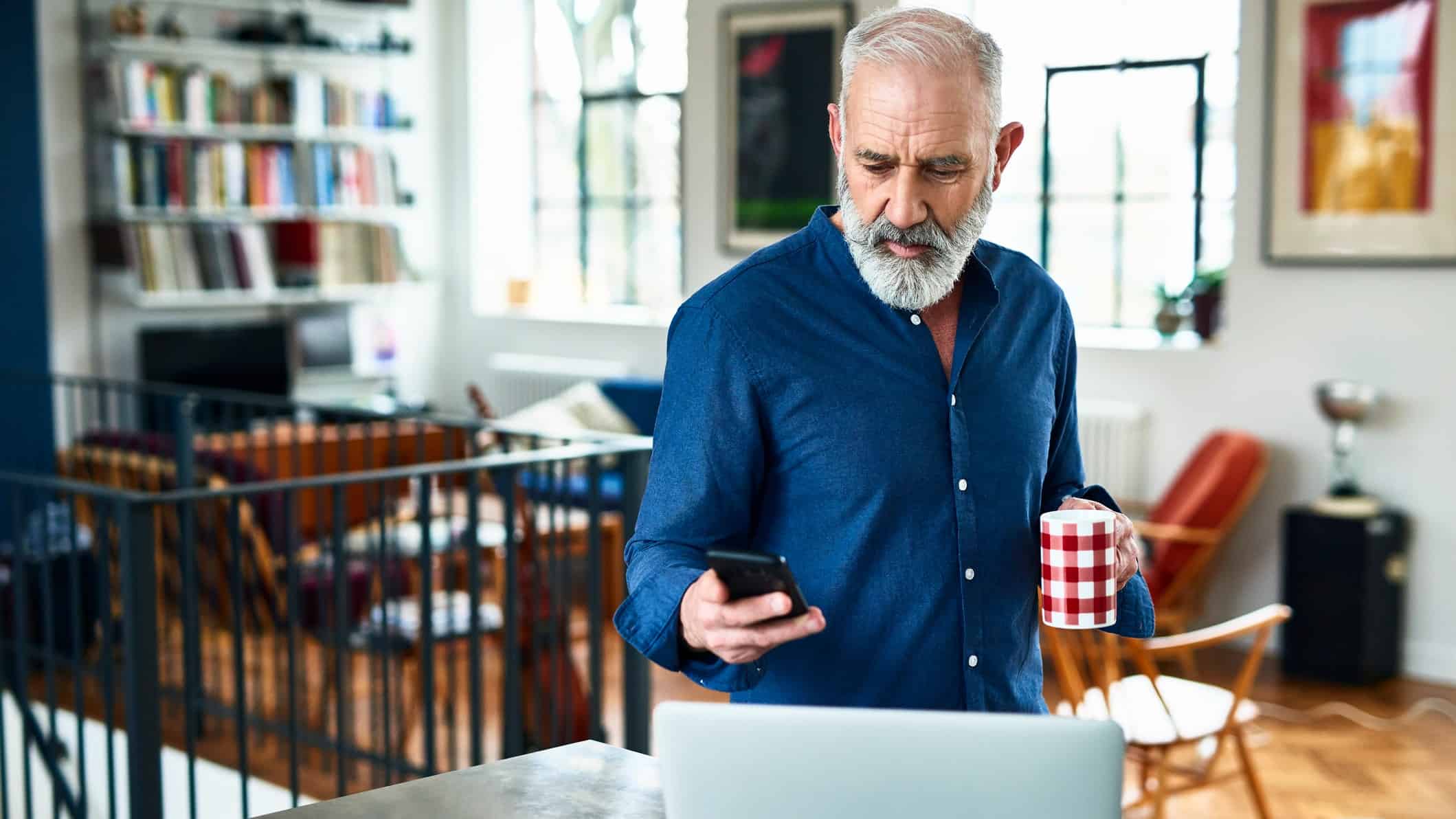 A middle aged man working from home looks at his iphone with a laptop open on the table in front of him