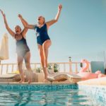Two elderly retired women jump into a pool together laughing.