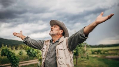 A farmer stands with arms outstretched to the sky as dark clouds billow overhead.