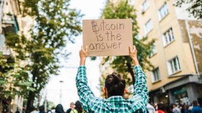 A protestor holds a cardboard sign saying, Bitcoin is the answer.