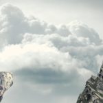 A boy standing on the edge of a cliff peers at a red flag in the distance through binoculars.