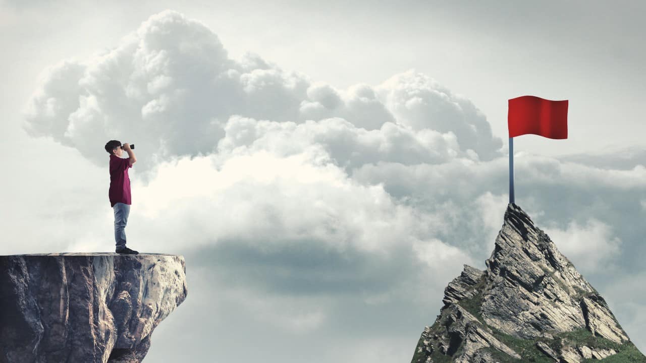 A boy standing on the edge of a cliff peers at a red flag in the distance through binoculars.