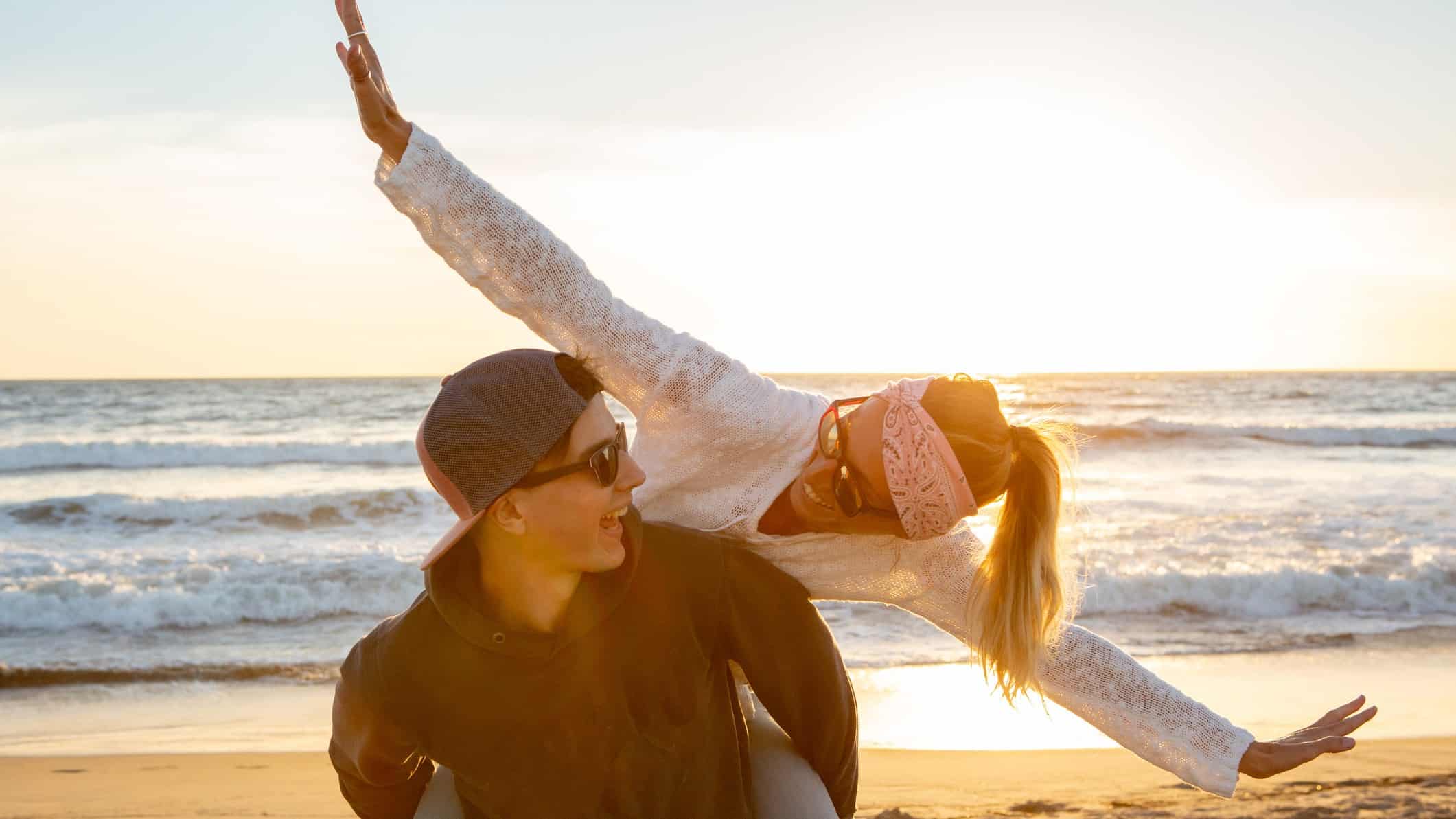 A happy woman flies with arms outstretched on her boyfriend's back on the beach at dusk.