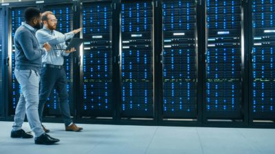 Two IT professionals walk along a wall of mainframes in a data centre discussing various things