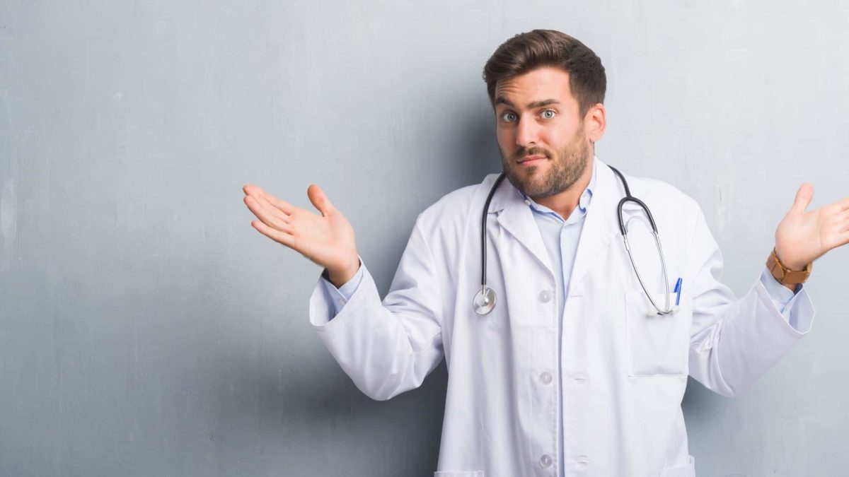 A male doctor wearing a white lab coat shrugs his shoulders and holds his hands up in the air looking confused