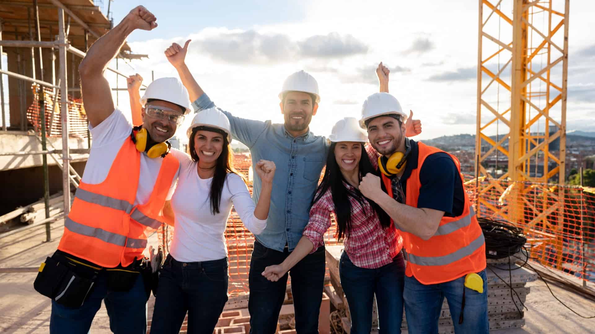 a group of five engineers wearing hard hats and some in high visibility vests raise their arms in happy celebration atop a building site with construction and equipment in the background.