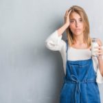 a woman stands with her hand to the side of her head and a sad, slightly distressed look to her expression while holding a large glass of milk in her other hand.