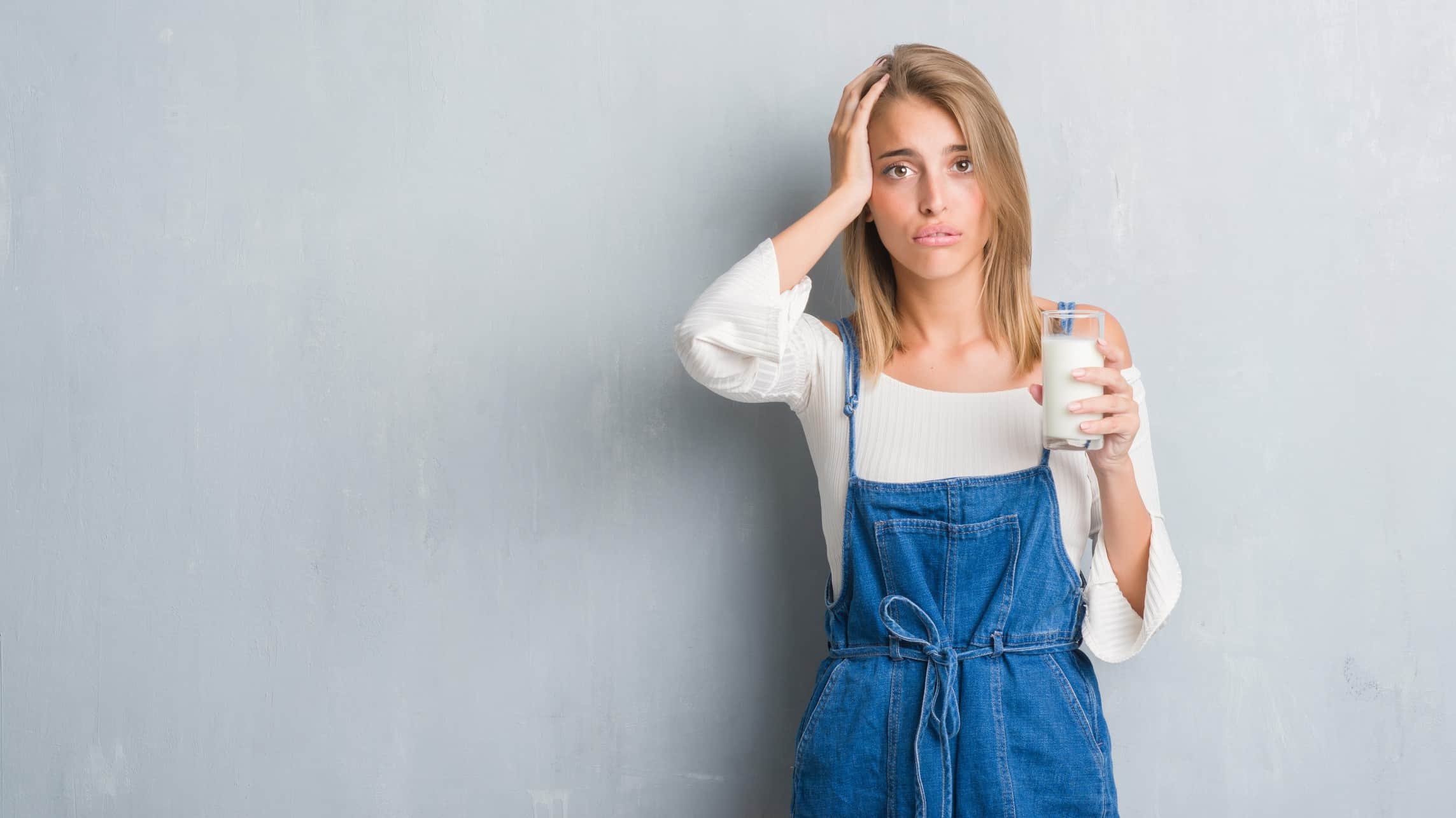 a woman stands with her hand to the side of her head and a sad, slightly distressed look to her expression while holding a large glass of milk in her other hand.