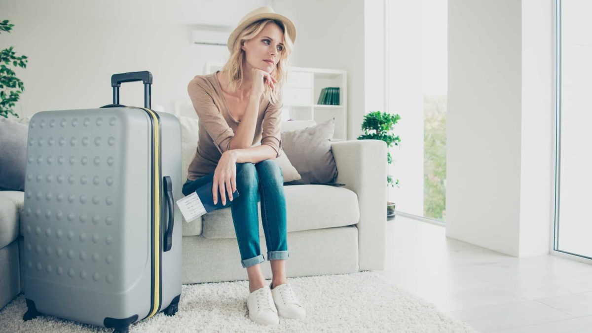 A pensive-looking woman sits on a chair with her chin on her hand looking into space with a large suitcase standing beside her as she contemplates travel to Europe and the Flight Centre share price