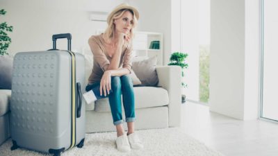 A pensive-looking woman sits on a chair with her chin on her hand looking into space with a large suitcase standing beside her as she contemplates travel to Europe and the Flight Centre share price