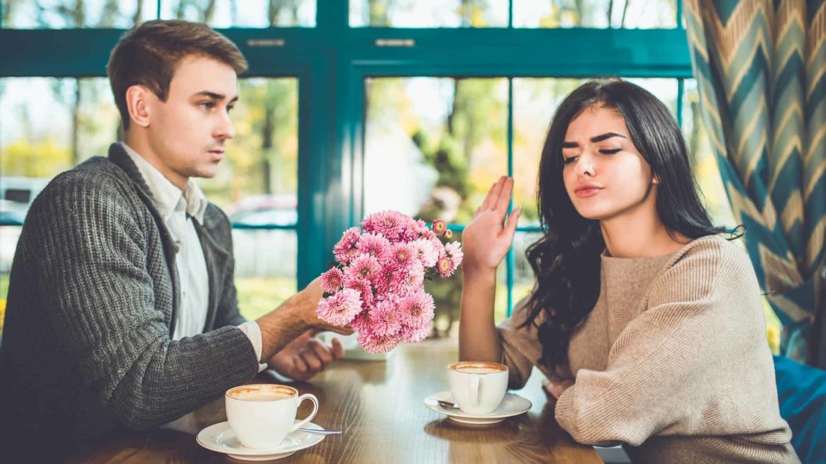 A woman holds up her hand in a stop gesture with a suspicious look on her face as a man sitting across from her at a cafe table offers her flowers.
