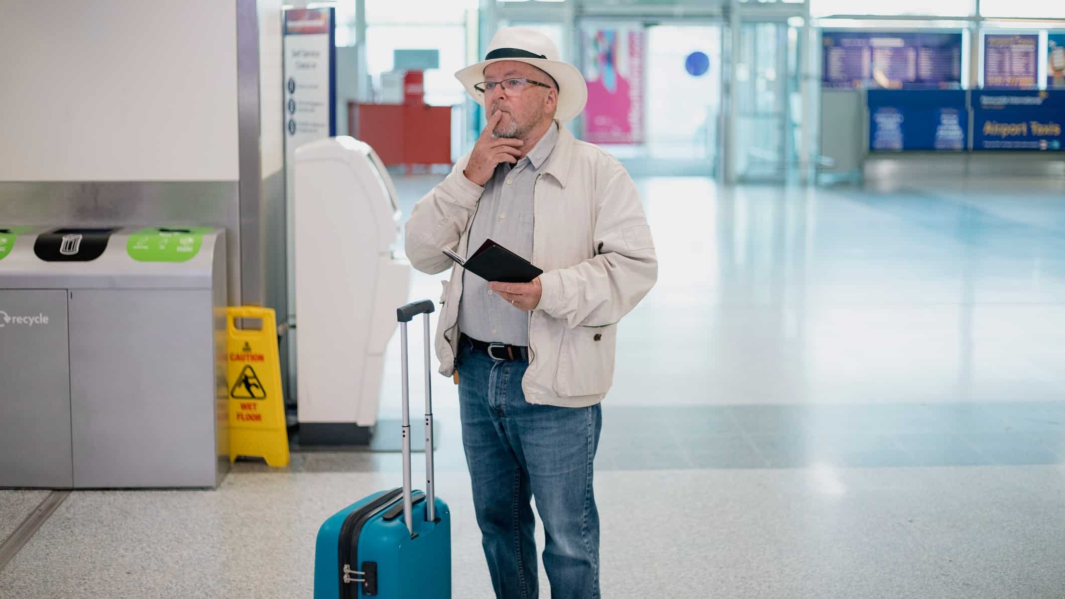 a man stands with travel documents in hand with a roller wheel suitcase and extended handle next to him holding his forefinger to his lip as he ponders his next move in a deserted airport.