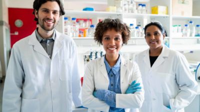 A group of medical researchers stands side by side with each other wearing white coats in their research laboratory with scientific equipment in the background.