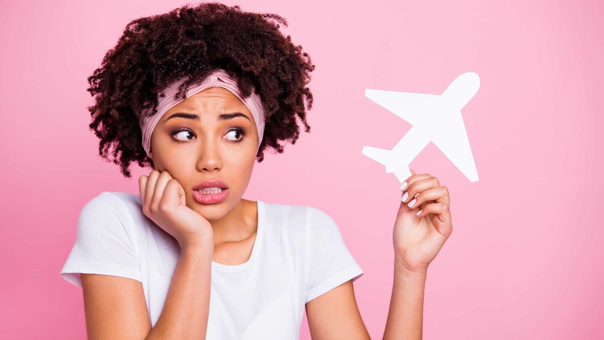 A woman looks nervous and uncertain holding a hand to her chin while looking at a paper cut out of a plane that she's holding in her other hand. representing the falling Air New Zealand share price today