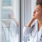 a doctor wearing a white coat with a stethoscope around her neck stares out a window with her hand to the side of her face as though in deep thought.