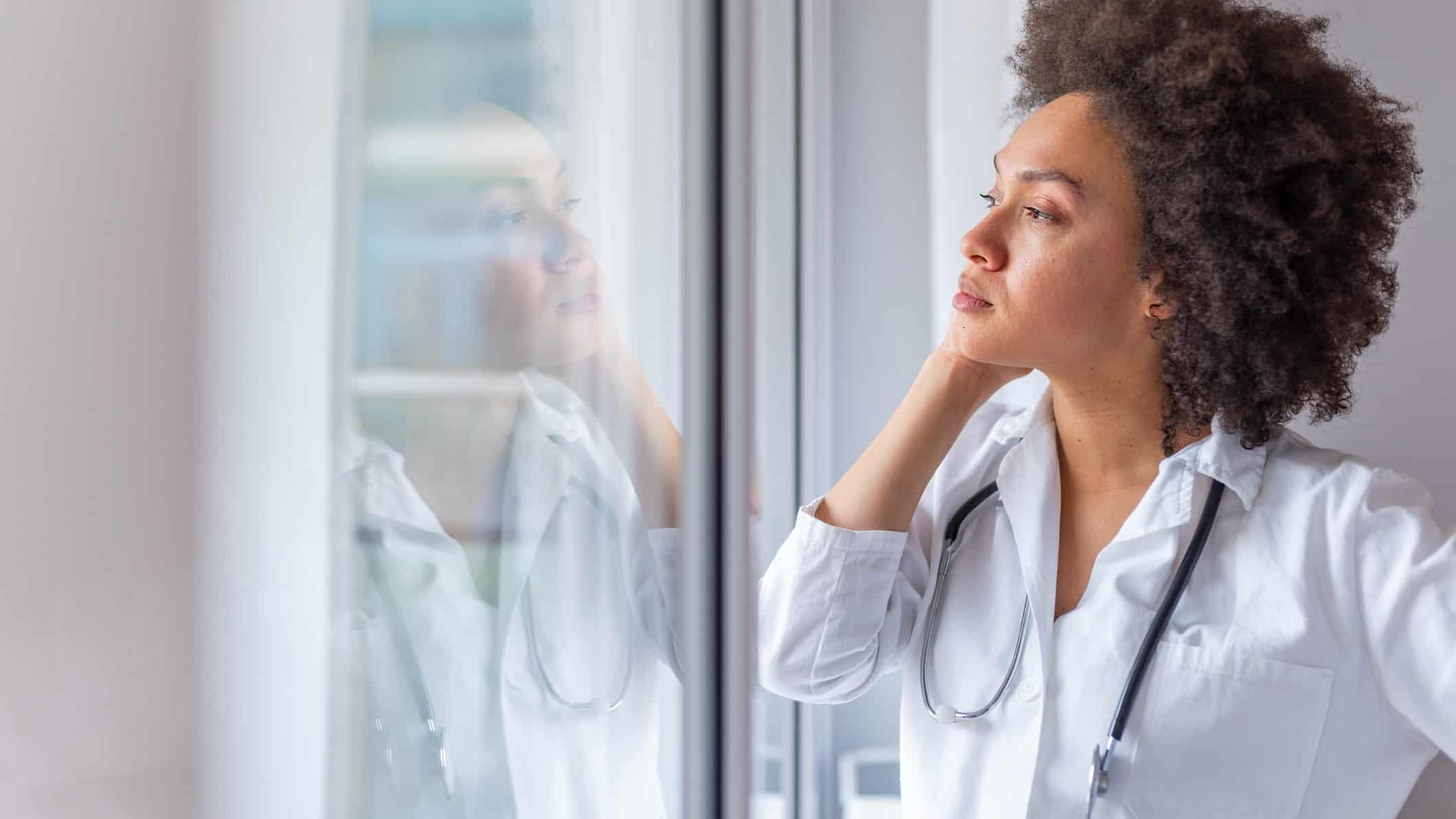 a doctor wearing a white coat with a stethoscope around her neck stares out a window with her hand to the side of her face as though in deep thought.