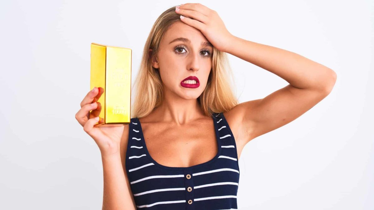 A woman holds a gold bar in one hand and puts her other hand to her forehead with an apprehensive and concerned expression on her face after watching the Ramelius share price fall today