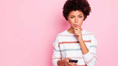 A woman with a mobile phone in her hand looks sceptical with a puzzled expression on her face with an eyebrow raised and pursed lips.