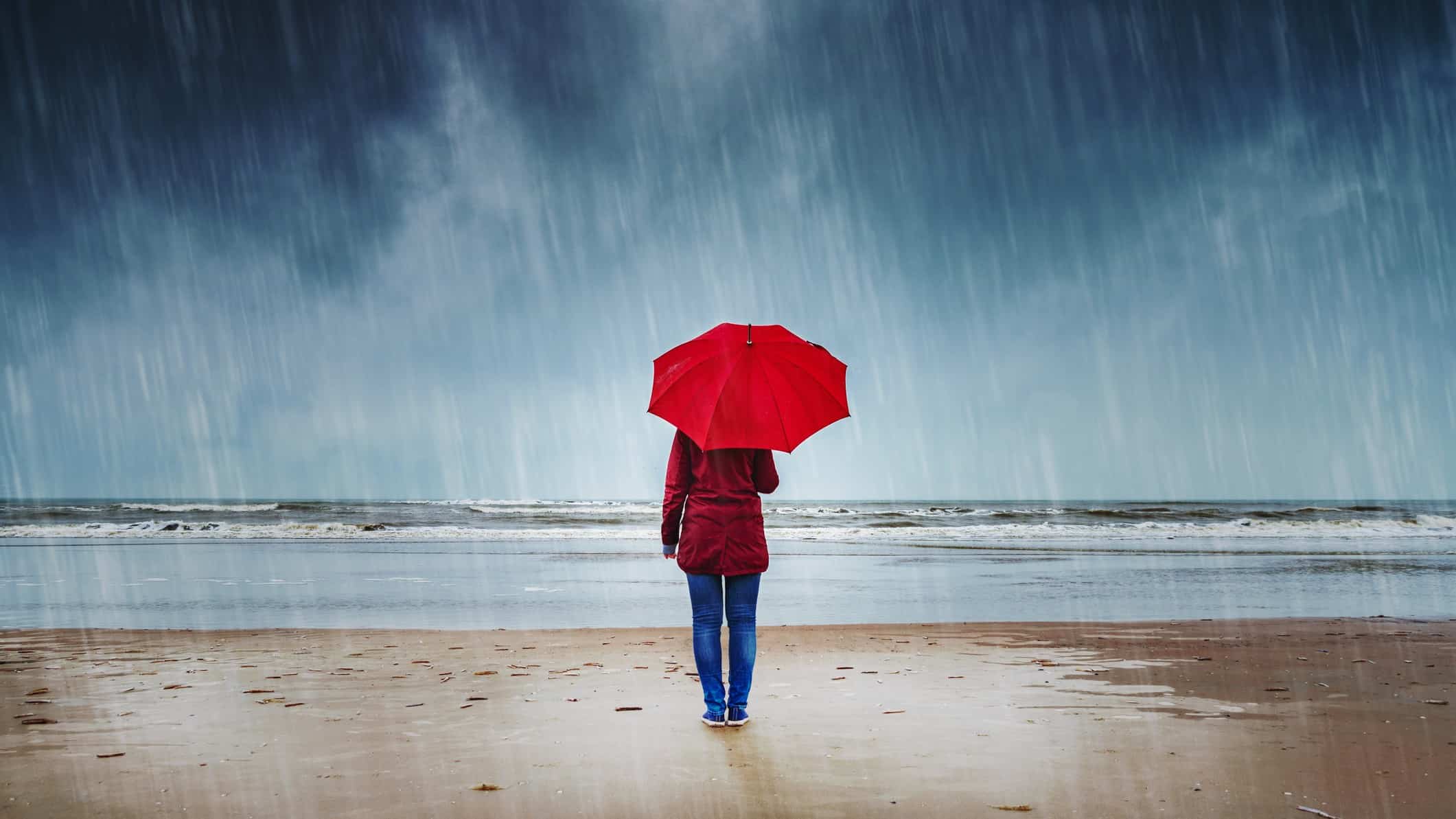 a woman under an umbrella stands looking out to sea on a beach in heavy rain with grey clouds gathering over the ocean.