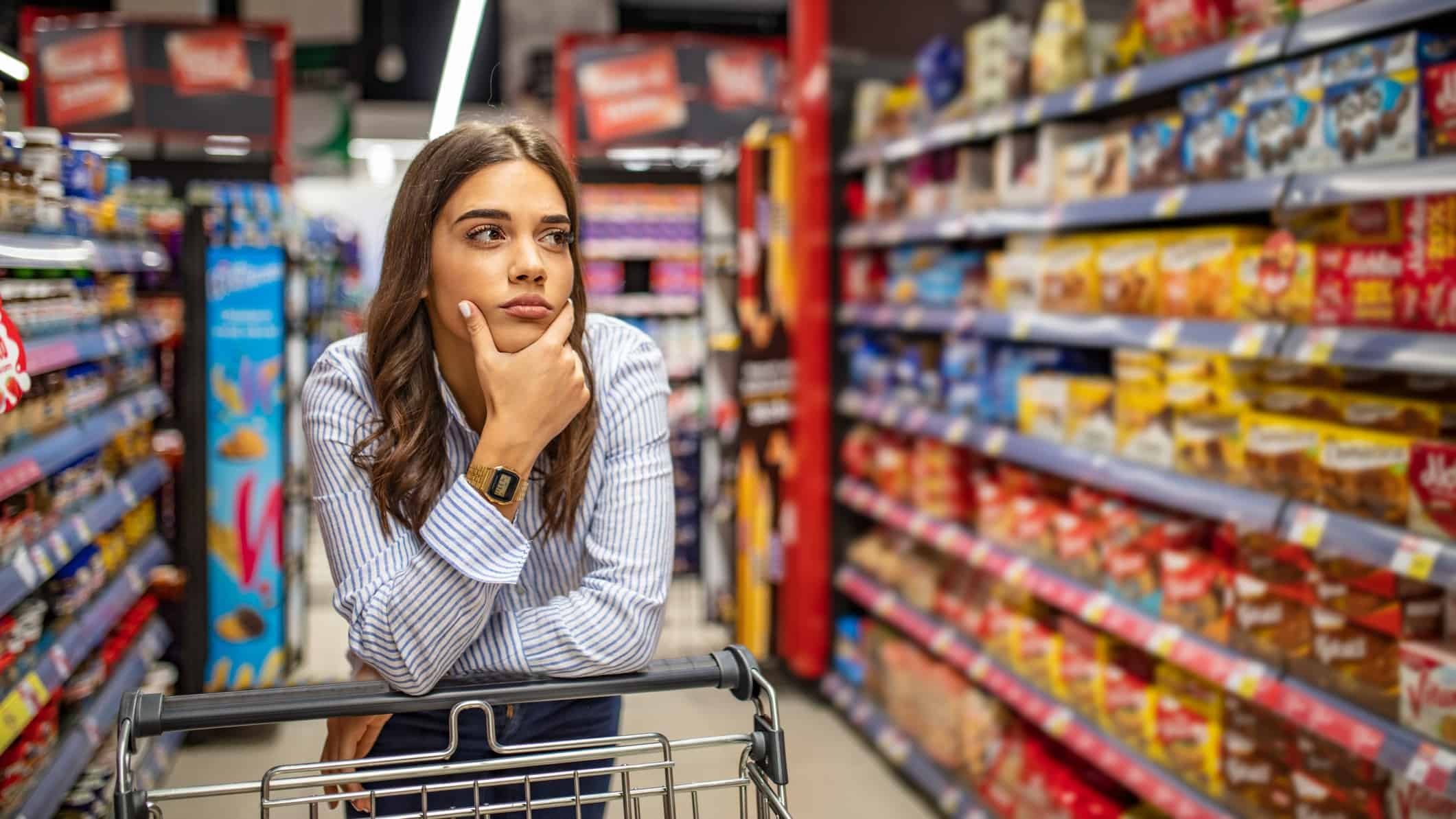a woman leans on her shopping trolley as she rests her chin in her hand as if thinking as she stands in the middle of a grocery supermarket shopping aisle with a serious look on her face.