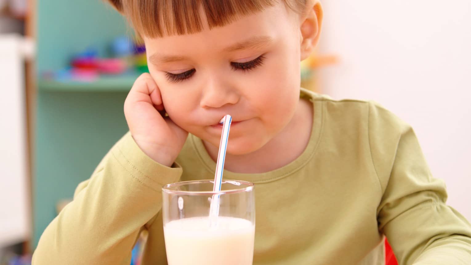 a small girl sits with her hand holding up the side of her face as she looks down in a downcast manner as she drinks a glass of milk through a straw.