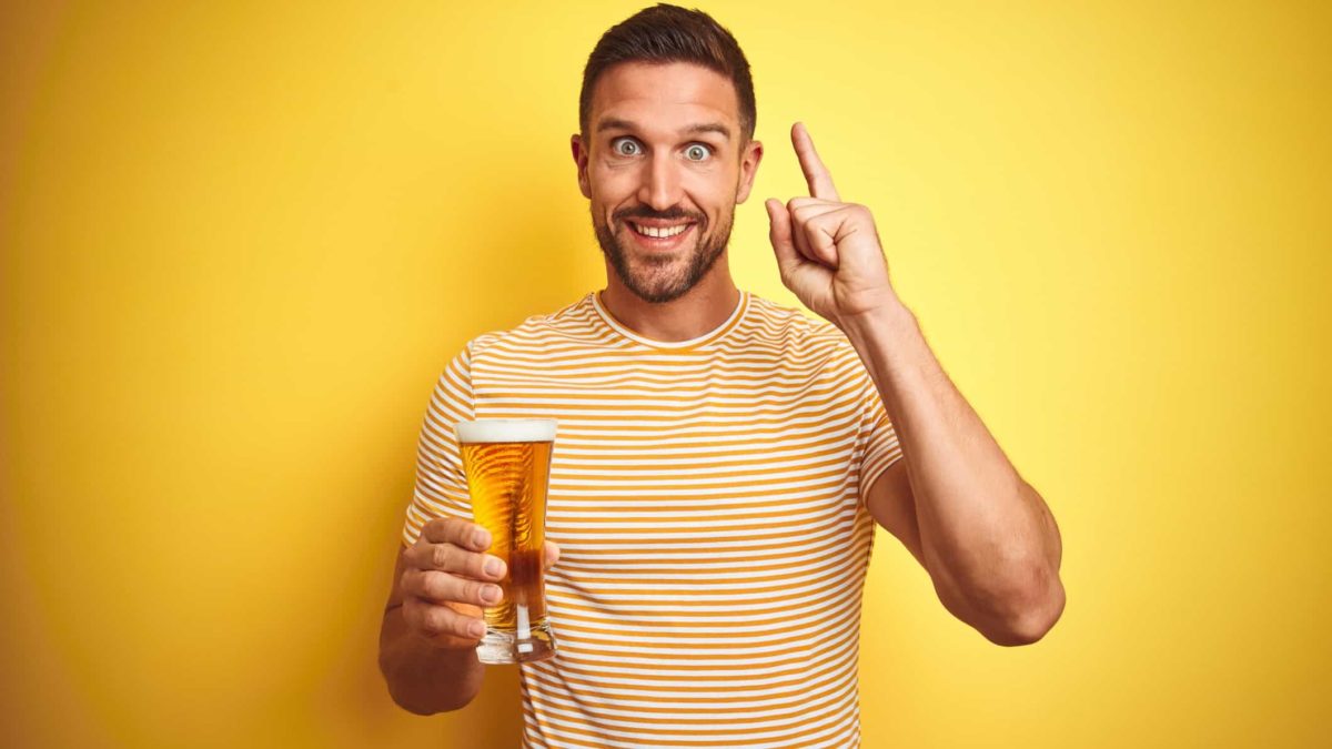 a man holding a glass of beer raises a finger with his other hand with a look of eager excitement on his face.