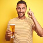 a man holding a glass of beer raises a finger with his other hand with a look of eager excitement on his face.
