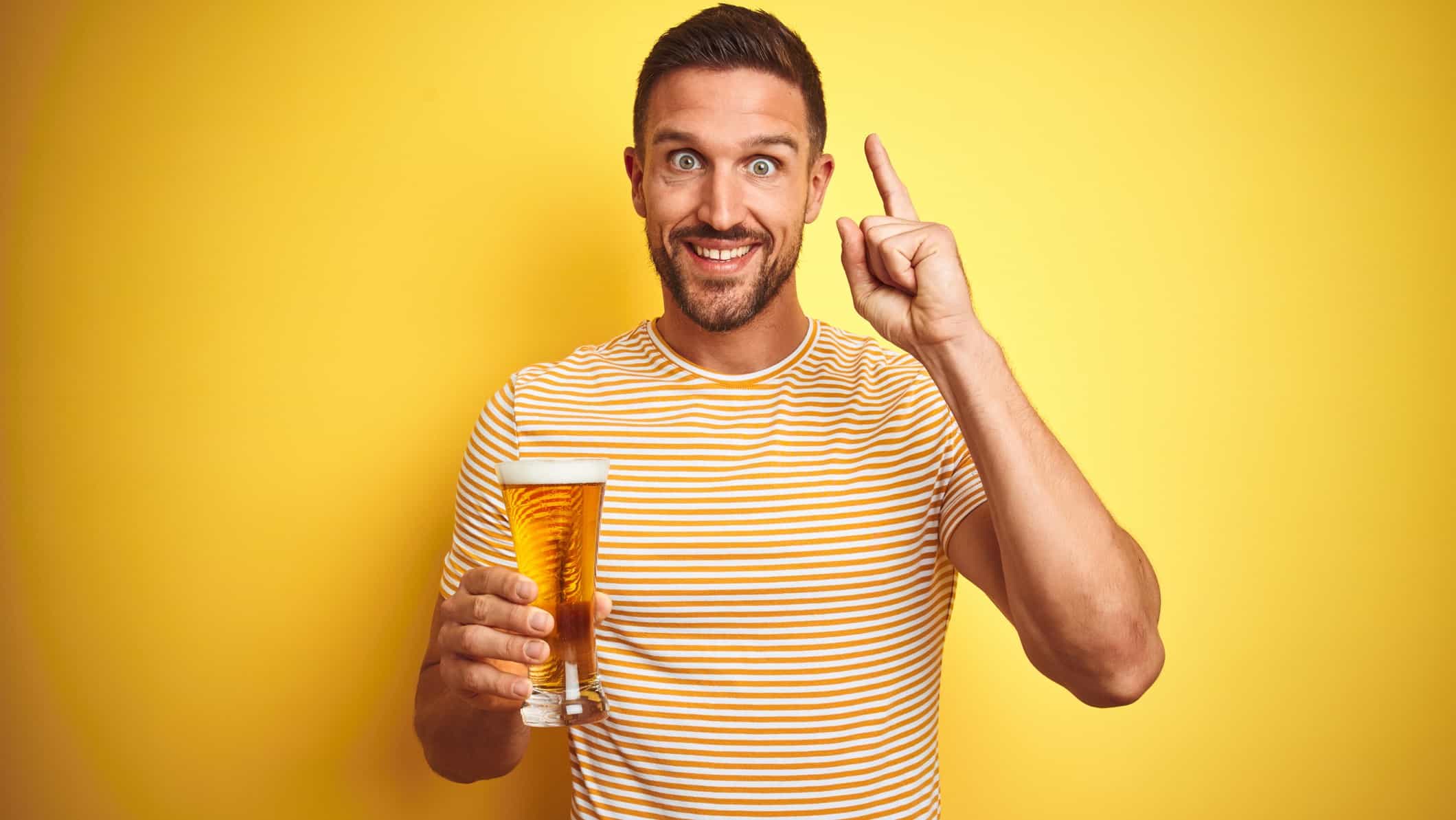 a man holding a glass of beer raises a finger with his other hand with a look of eager excitement on his face.
