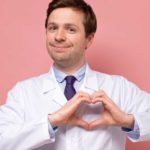 a doctor in a white coat makes a heart shape with his hands and holds it over his chest where his heart is placed.