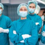 a group of surgeons in full surgery dress including masks, gloves and head coverings stands together with arms folded and smiling eyes as if happy with the outcome of their efforts.