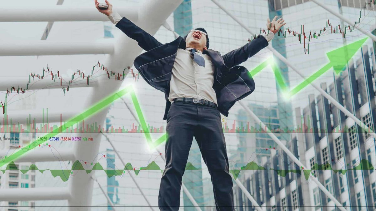 a man in a business suite throws his arms open wide above his head and raises his face with his mouth open in celebration in front of a background of an illuminated board tracking stock market movements.