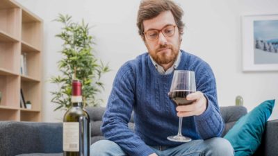 a man sits alone in his house with a dejected look on his face as he looks at a glass of red wine he is holding in his hand with an open bottle on the table in front of him.
