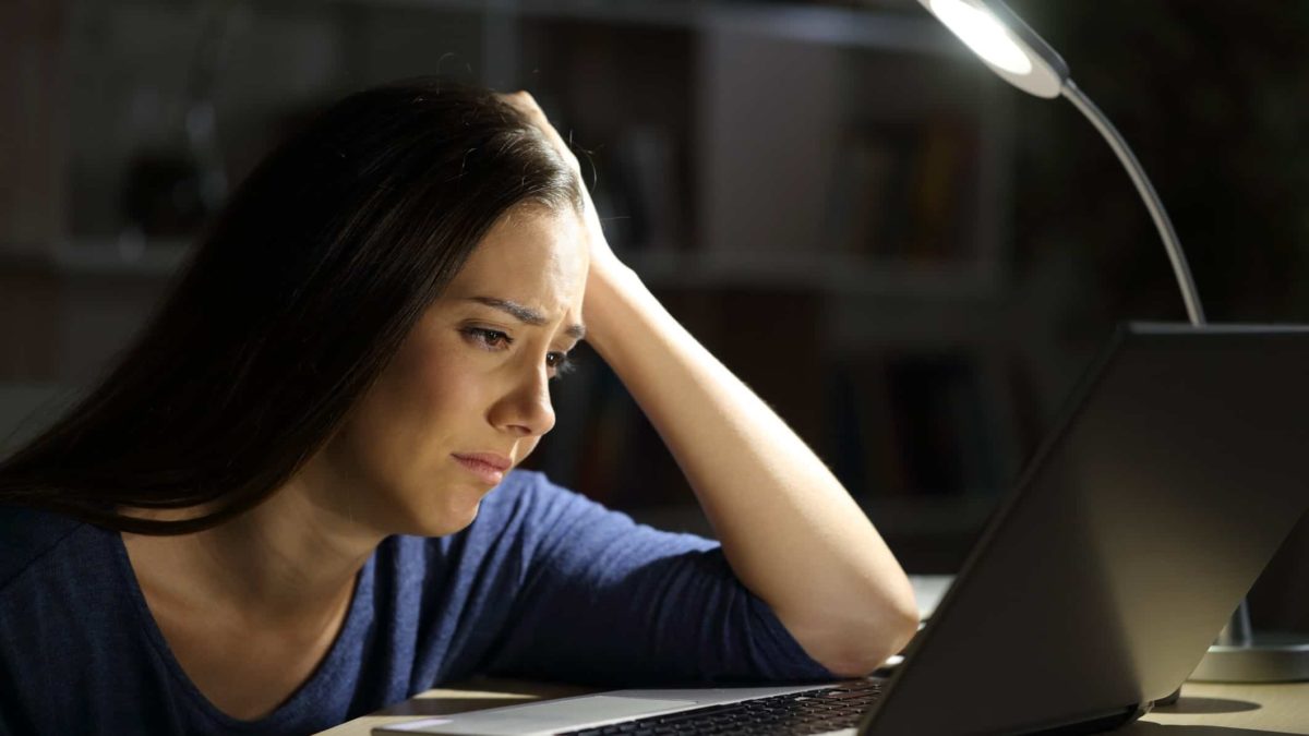 a woman sits with a sad and pained expression on her face as she slumps over her laptop computer on a desk with a desk lamp in the background.