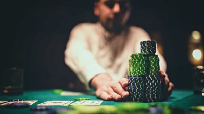 a person in the dark background of a casino gambling room places his hands either side of a large pile of casino chips on a card table.