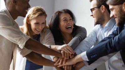 a group of business people in business attire join their hands in the middle of a circle in a team celebration as they smile broadly in celebration of a milestone event.