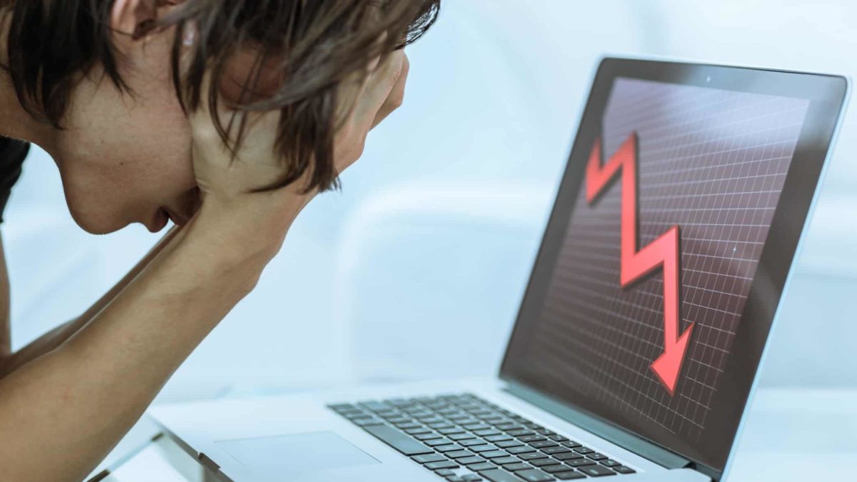 a person holds their head in their hands as they slump forward over a laptop computer which features a thick red downward arrow zigzagging downwards across the screen.