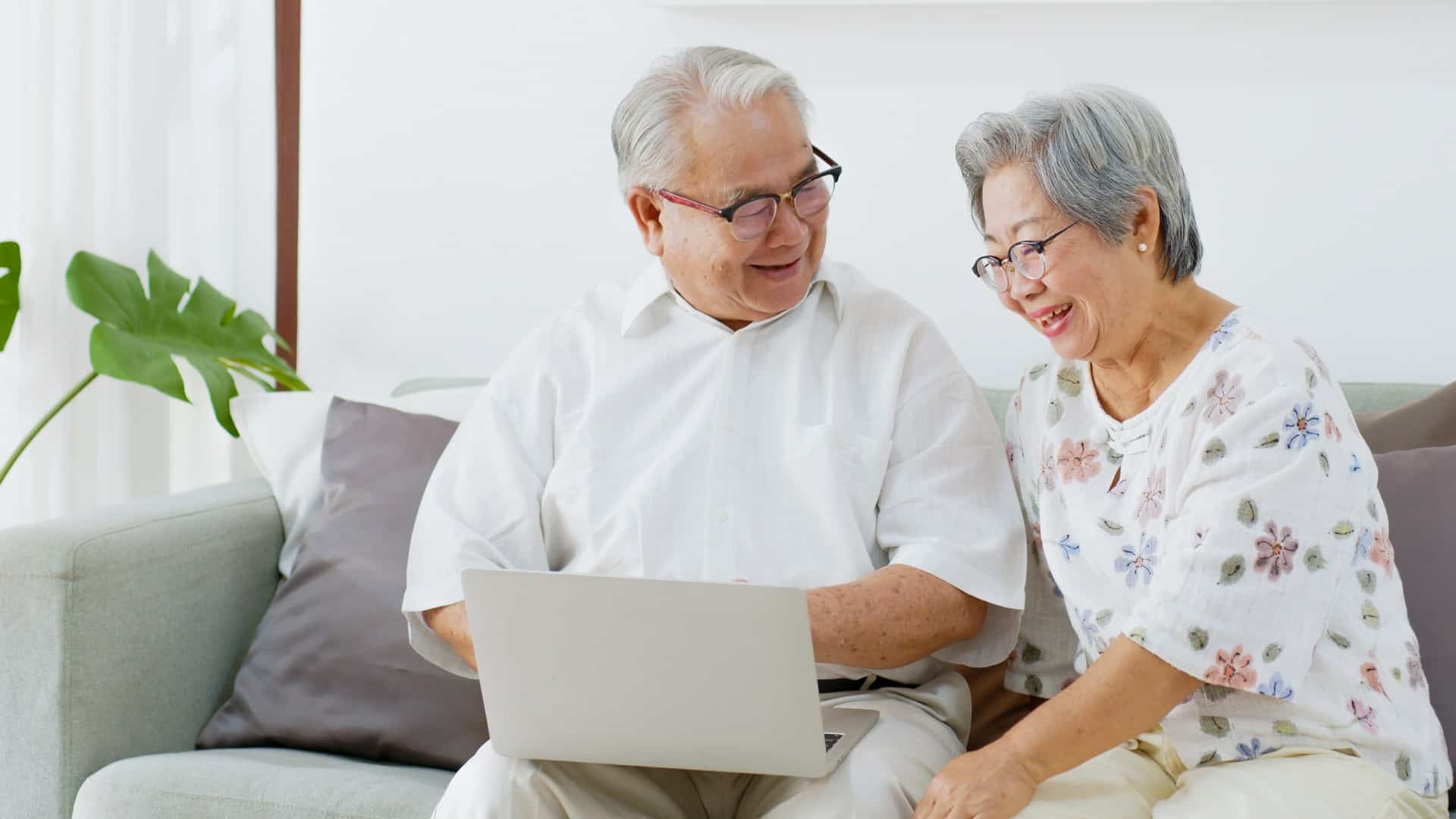 an older couple look happy as they sit at a laptop computer in their home.