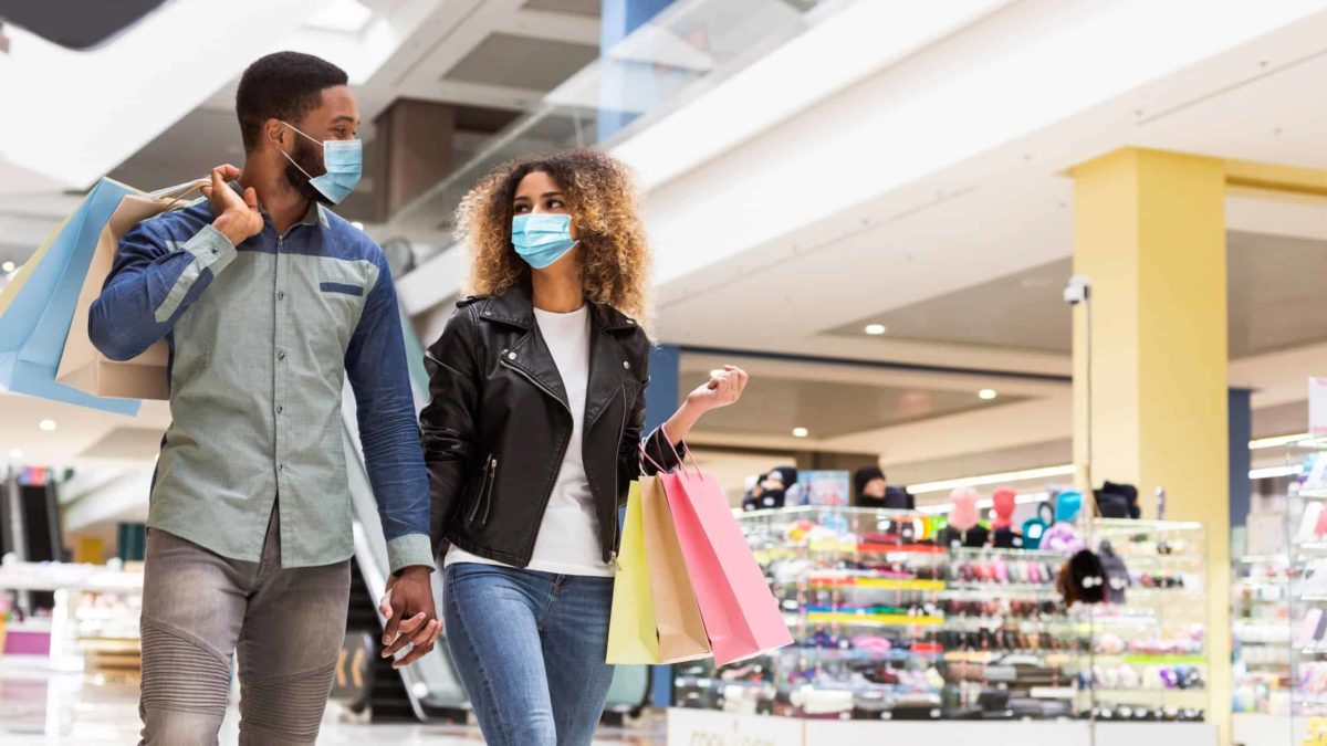 a man and a woman hold hands wearing masks as they carry shopping bags and stroll through a retail shopping centre.