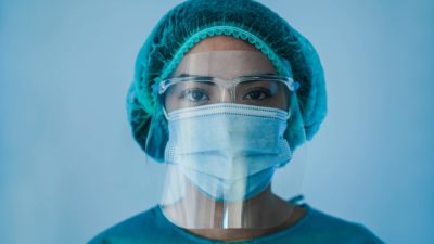a healthcare worker wears full personal protective equipment including a face shield, mask, head covering and gown.