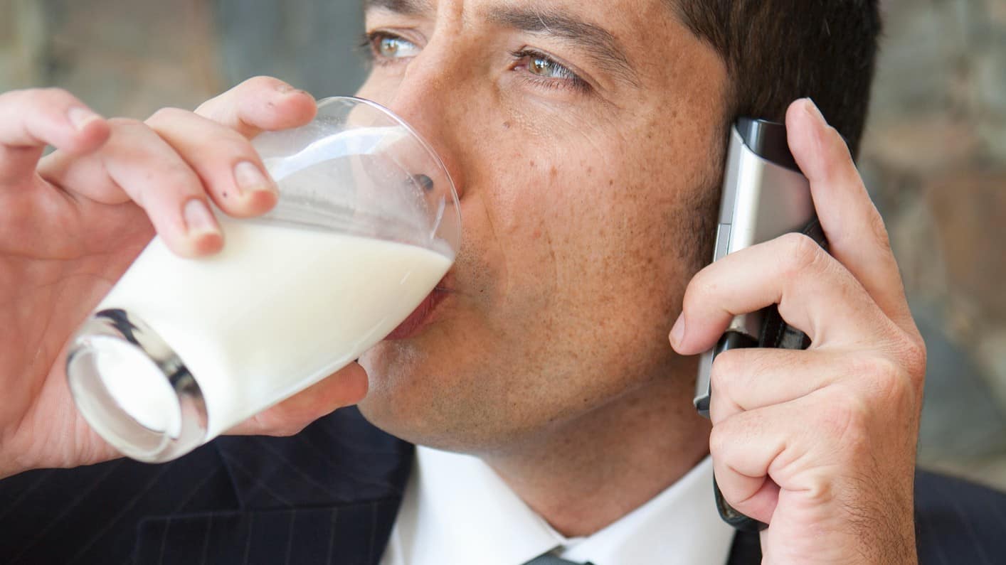 a man in a business shirt, tie and suit holds a mobile phone to his ear while he drinks a large glass of milk.