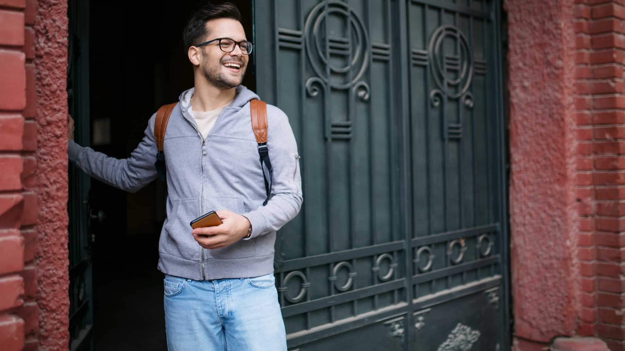 a man in casual clothes emerges from his front door carrying a phone and wearing a backpack with a wide smile on his face.