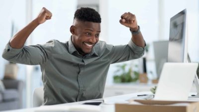 a man raises his fists to the air in joyous celebration while learning some exciting good news via his computer screen in an office setting.