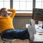 a bearded man sits at his desk with hands behind his head and feet on his desk smiling widely while looking at his computer screen which has market data on it, indicating a please share price rise.