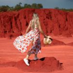 a woman in a flowing dress stands against the backdrop of red iron ore rich dirt as in central Australia.