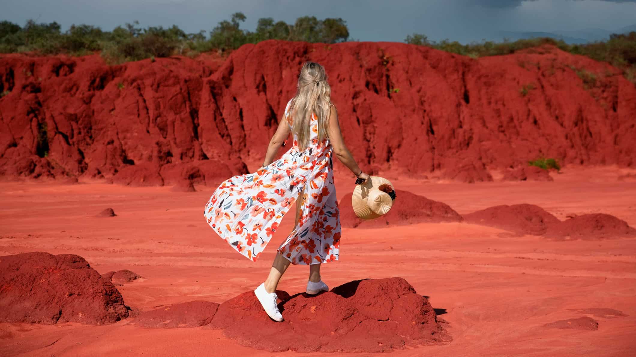 a woman in a flowing dress stands against the backdrop of red iron ore rich dirt as in central Australia.