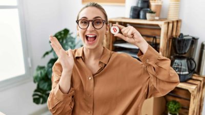 A young woman wearing work wear in an office setting has a lively, happy, open-mouthed expression of joy while holding a bitcoin token.