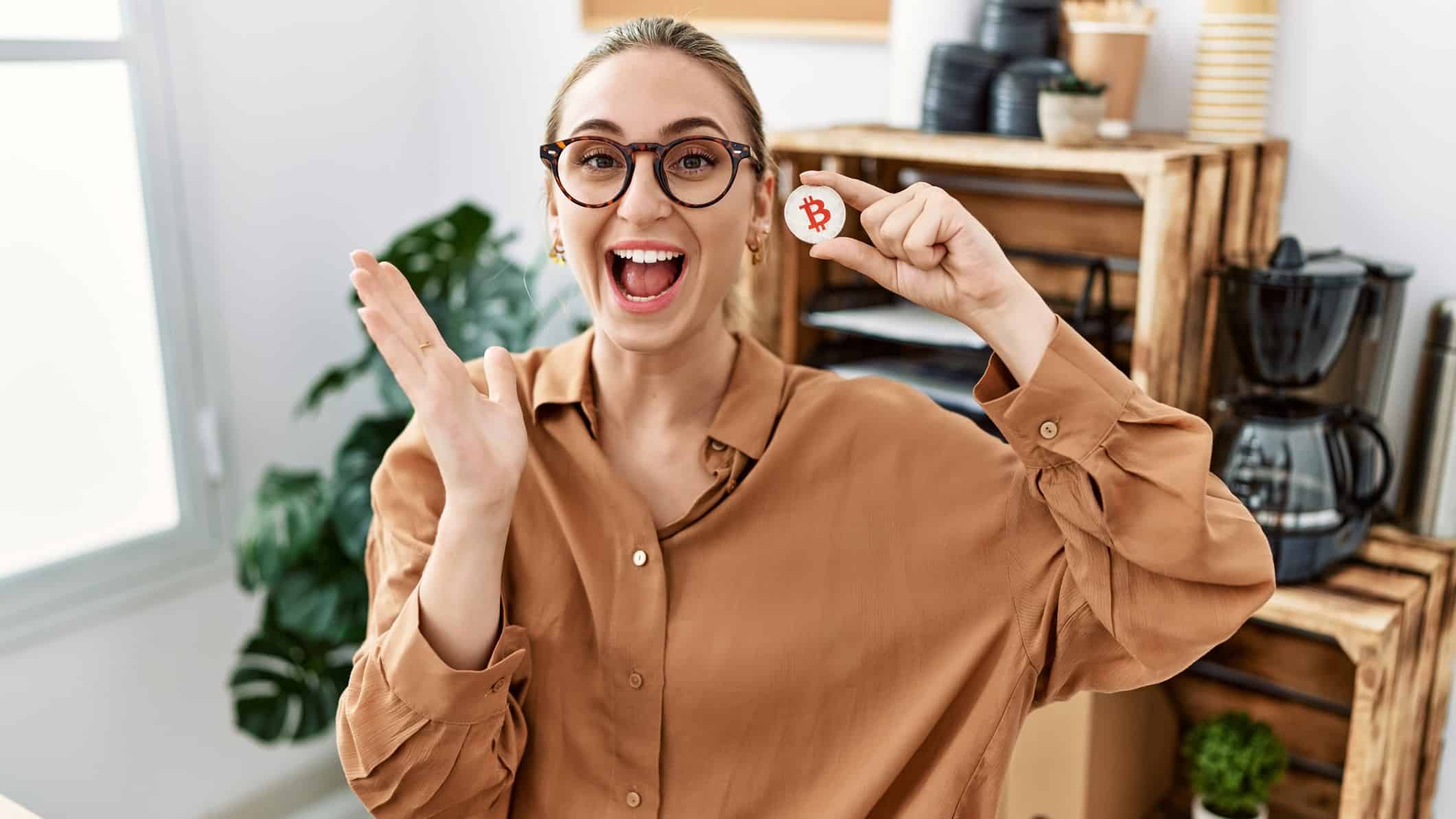 a young woman wearing work wear in an office setting has a lively, happy openmouthed expression of joy while holding one hand up in a happy gesture while holding a bitcoin token in the other hand.