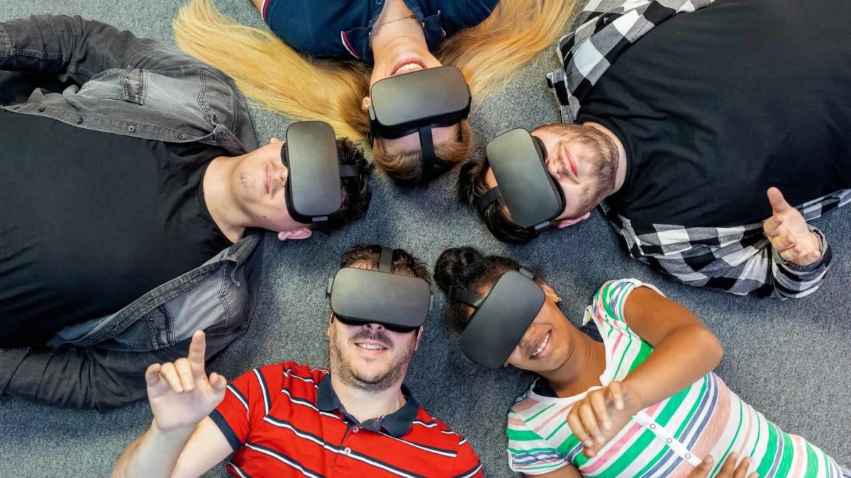 a group of five people lie on the floor with their heads touching, each wearing hi tech goggles over their eyes as if in a metaverse workplace collaboration.
