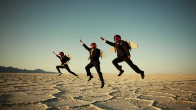 three young children weariing business suits, helmets and old fashioned aviator goggles wear aeroplane wings on their backs and jump with one arm outstretched into the air in an arid, sandy landscape.