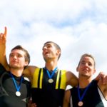 three men stand on a winner's podium with medals around their necks with their hands raised in triumph.
